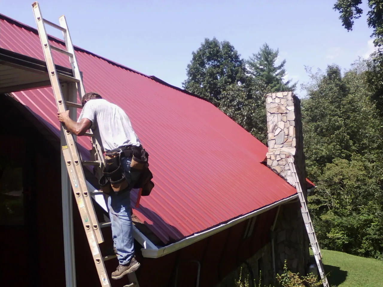 Roofer on a ladder beside a red metal roof and stone chimney — Soffit & Fascia Repair by Roofing Brothers in Powers